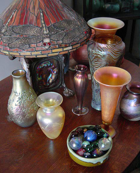 An Art Glass Collection Featuring a Dragonfly Stained Glass Lamp Plus Art Glass Vases and Marbles: : This stained glass lamp is filling double roles in the room. It is useful accent lighting that adds shimmery luminescence to the collection of art glass vases surrounding it. With its colorful stained glass lampshade and graceful form, it also becomes an integral part of the art glass collection. The dragonfly motifs draw the eye to create a focus of interest, while the bronze lamp base adds an interesting counterpoint to all the shimmering glass in the collection.